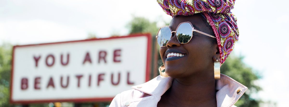 Smiling woman stands in front of a sign saying 'you are beautiful'