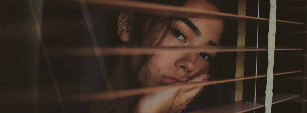 Woman stares out through window blinds, looking bored