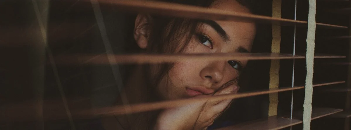 Woman stares out through window blinds, looking bored